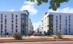 Modern white apartment buildings flank a wide pedestrian plaza with trees under blue sky