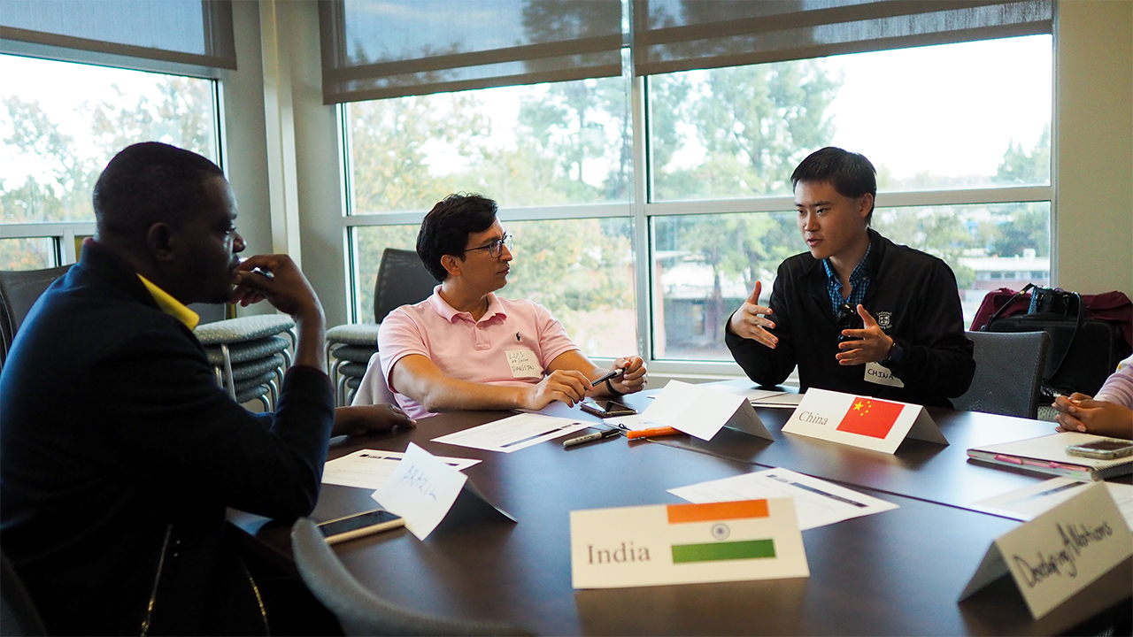 Silas sits in profile at a work table with Luis at his left. They both look at and listen to a male student representing China at the right of the photo, who is speaking and gesturing conversationally with his hands. Placards on their table identify the group as \"Developing A Nations\" and participants represent countries including China, India and Brazil.