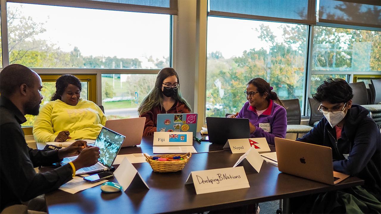 Gathered around a table with windows behind them looking out on trees and a grassy field, Edu Nguema, Rudo Chasi, a female student in a mask, Sushila Thing and a male student in a mask each have a laptop in front of them. Placards on the table identify this group as \"Developing B Nations\" and some of the countries they represent as Fiji and Nepal.
