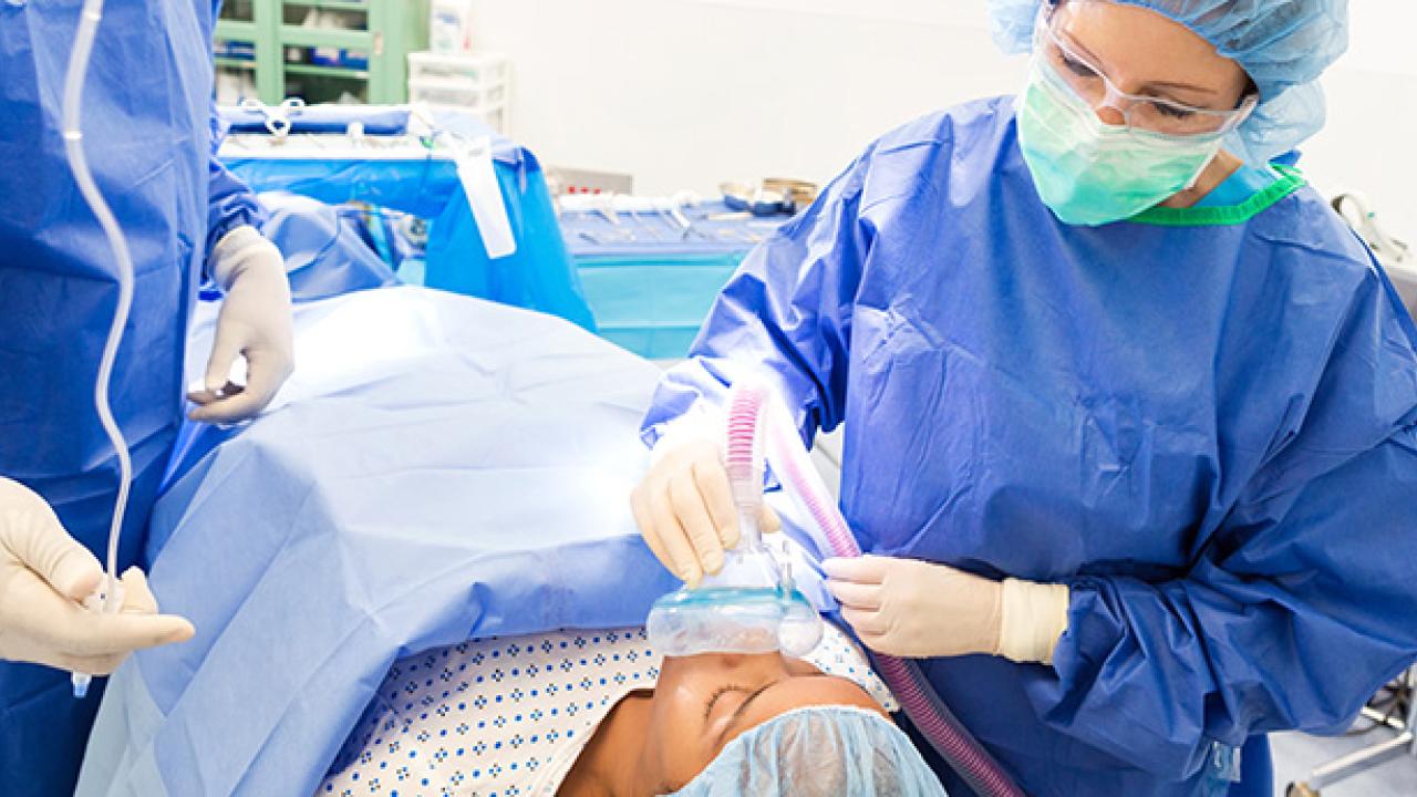 A young patient is being sedated while laying on operating table. A female anesthesiologist is using medical equipment to sedate patient.