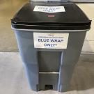 Gray plastic bin with black lid labeled "BLUE WRAP ONLY" on a concrete floor.